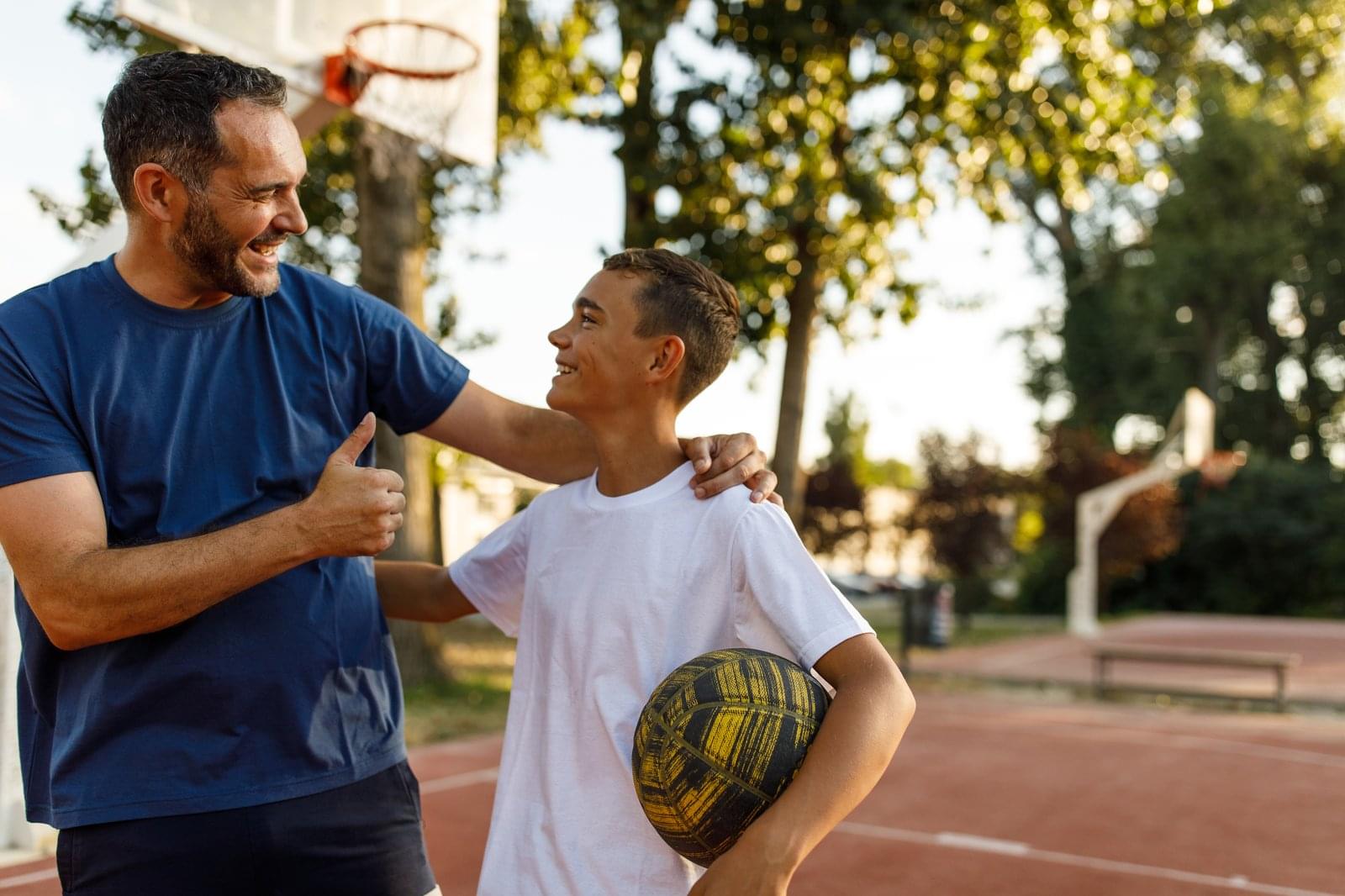 Smiling father giving his teenage son a thumbs-up on a basketball court at sunset